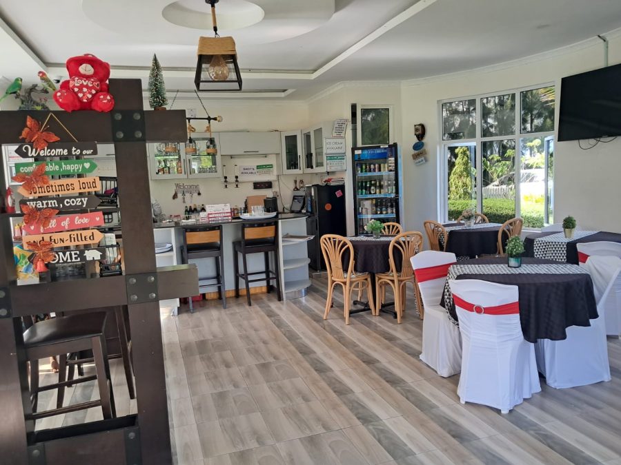 The interior restaurant and bar area of Olunana Gardens on Kangundo Road. The space features modern wood-look flooring, large windows with garden views, and a mix of dining tables with black and white linens. A well-stocked bar with high stools and a beverage cooler are visible in the background, along with a decorative wooden partition and a wall-mounted TV
