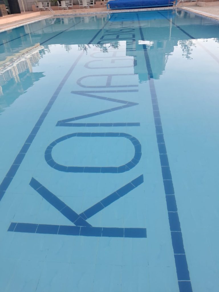 A large, clear swimming pool at Koma Gardens Resort with the brand name "KOMA GARDENS" tiled in bold blue letters on the pool floor, reflecting the sky and surrounding buildings.