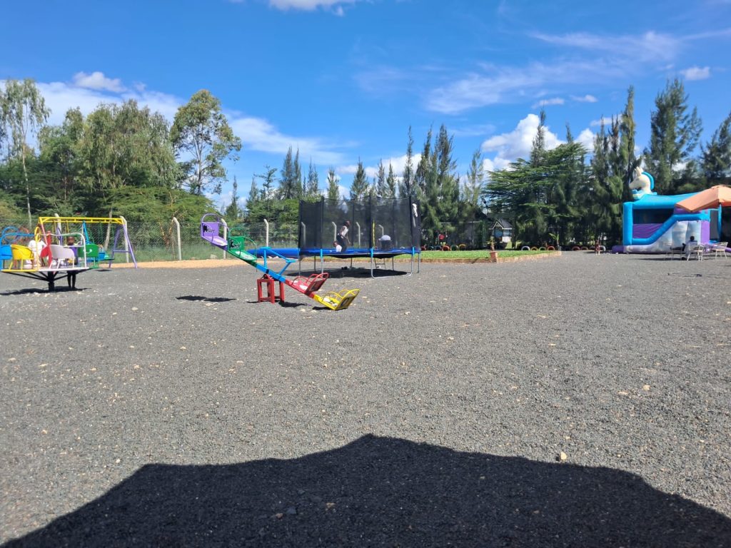 The expansive children's playground at Olunana Gardens on Kangundo Road. The image shows various play equipment on a gravel surface, including a colorful merry-go-round, a blue and red seesaw, an enclosed safety trampoline, and a large blue bouncy castle in the background under a clear sky.