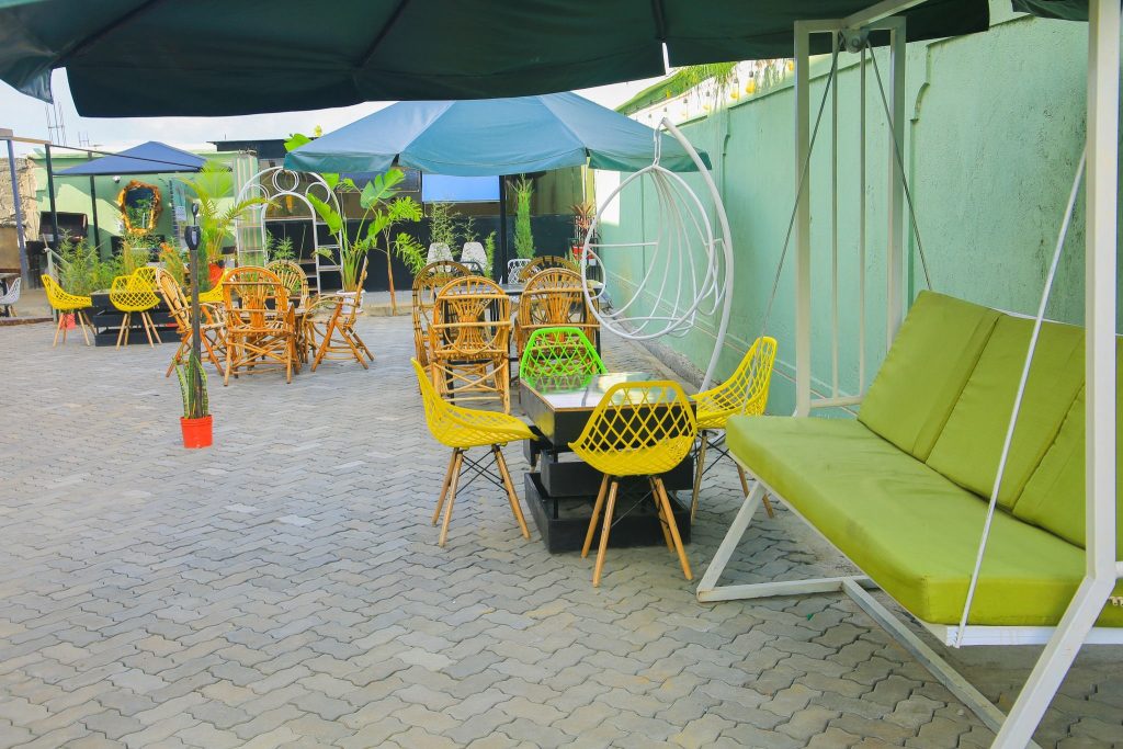 Outdoor patio seating area with gray brick paving. Features include bright yellow molded chairs around a table, a white metal swing couch with lime green cushions, a hanging egg chair, and traditional bamboo chairs under large dark green umbrellas against a mint green wall.