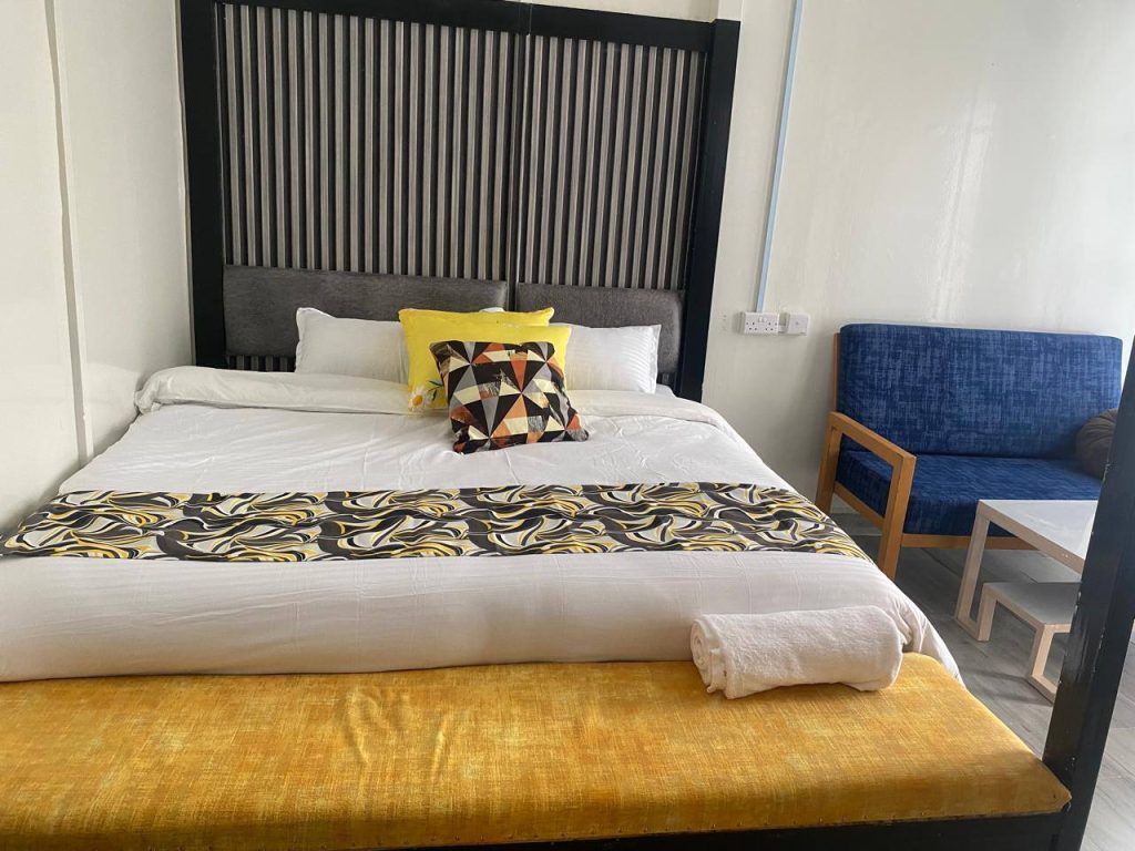 An interior view of a modern guest bedroom at Micaphonia Suites in Joska, featuring a large bed with a tall, black textured headboard, white linens, yellow decorative elements (bench and pillow), and a blue upholstered armchair with a wooden frame and side tables.